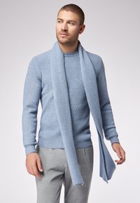 Uomo che indossa un maglione a coste blu chiaro con una sciarpa abbinata drappeggiata attorno al collo, abbinato a pantaloni grigi a quadri, che guarda verso sinistra.