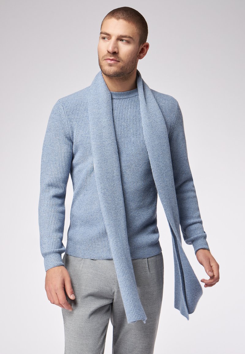Uomo che indossa un maglione a coste blu chiaro con una sciarpa abbinata drappeggiata attorno al collo, abbinato a pantaloni grigi a quadri, che guarda verso sinistra.