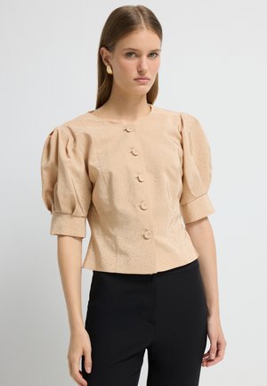 Luchtige blouse met opgezette mouwen in lichtbeige met subtiele metallic spikkels, voorzien van een ronde halslijn en zes knopen. Gstyled met zwarte broeken.