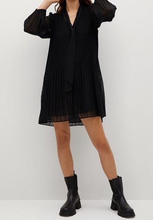 Robe noire plissée avec un col noué, manches transparentes et texture à pois, associée à des bottines noires épaisses.