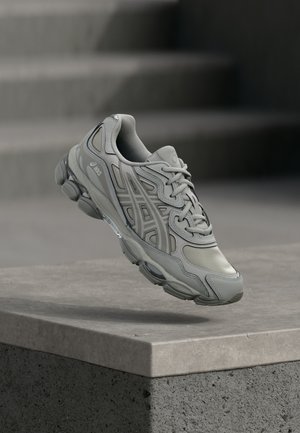 Szary but do biegania Asics z detalicznym wzorem unoszący się nad betonowym krawężnikiem, w tle rozmyte schody.