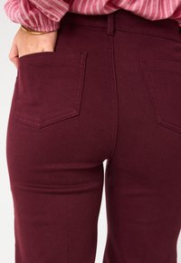 Pantaloni in denim bordeaux con vita alta, taglio dritto e tasche posteriori. Il tessuto appare liscio con dettagli di cucitura sottili.