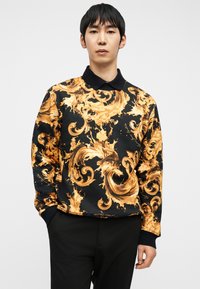 Άνδρας που φοράει μαύρο φούτερ με έντονα χρυσά μπαρόκ στροβιλισμένα μοτίβα και μαύρο γιακά, στέκεται με το ένα χέρι στην τσέπη μπροστά σε λευκό φόντο.