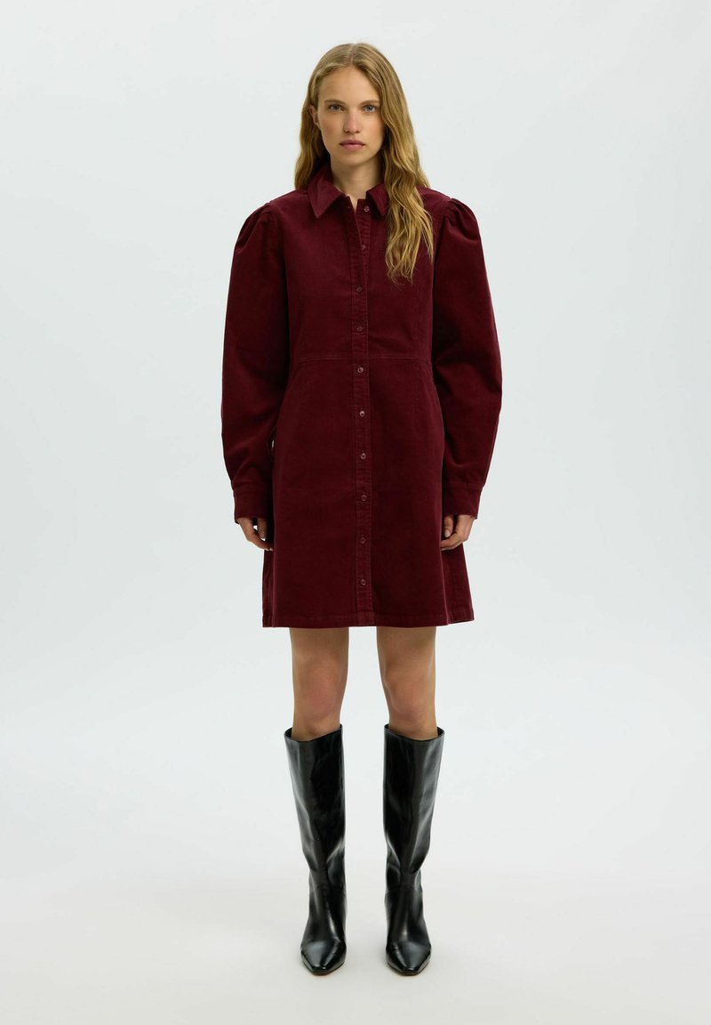 Femme aux longs cheveux ondulés portant une robe bordeaux boutonnée avec des manches bouffantes et des bottes noires montantes jusqu'aux genoux, debout sur un fond blanc.