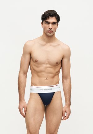Hombre musculoso con calzoncillos Dsquared2 de marca en azul marino y blanco, de pie contra un fondo claro y llano, mirando hacia adelante.