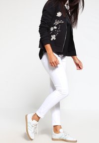 Sort bomberjakke med blomsterbroderi, hvide skinny jeans og hvide sneakers med lyseblå striber og beige sål.