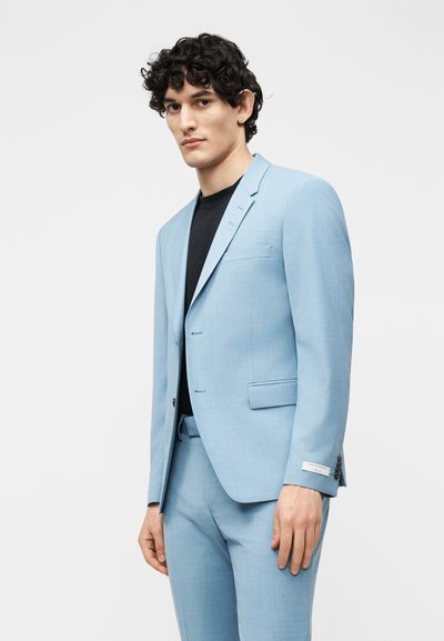 Νεαρός άνδρας με σγουρά μαλλιά, που φοράει γαλάζιο κοστούμι και μαύρο πουκάμισο, στέκεται μπροστά σε λευκό και απλό φόντο.