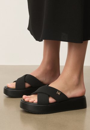 Fødder iført sorte plateau-slider-sandaler med brede krydsede stofstropper og en lille gulddetalje med logo på en neutral gulvflade.