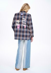 Kostičkovaný blazer v modré, hnědé a krémové barvě, s barevnými nášivkami vzadu. Vytvořený v kombinaci s světle modrými širokými džíny a hnědými botami.