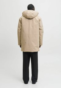 Bežna vodoodporna jakna z velikim kapuco, brezšivno zasnovo in ravnim krojem, v kombinaciji s črnimi hlačami in temnimi čevlji.