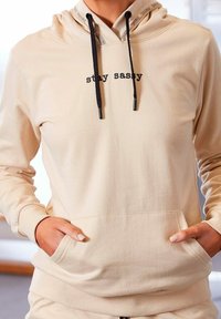 Sudadera con capucha beige con cordones negros, bolsillo tipo canguro y texto "stay sassy" en el frente. Tejido suave, ajuste cómodo y puños acanalados.