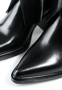 Bottes cheville en cuir noir brillant à bout pointu avec talons bas sur une surface blanche.