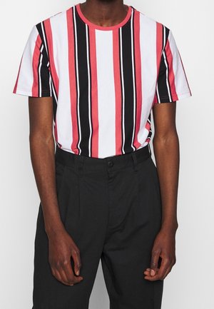 Homme portant une chemise à manches courtes avec des rayures verticales rouges, blanches et noires rentrée dans un pantalon noir taille haute.