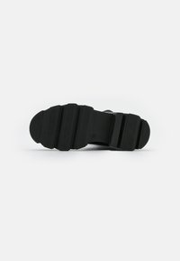 Suela de zapato negra con una superficie de goma texturizada que presenta múltiples surcos y crestas para mayor tracción. Forma de punta curvada.