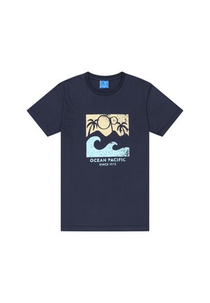 Homme avec des cheveux tressés et des tatouages aux bras, portant un t-shirt bleu marine avec un imprimé vague et palmier, un jean ample bleu clair et des baskets blanches.