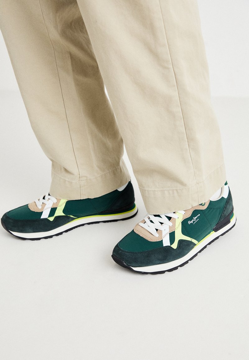 Zapatillas verdes y negras con materiales de ante y malla, cordones blancos, acentos amarillos y un diseño retro, combinadas con pantalones beige.