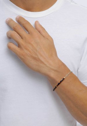 Main avec un bracelet à perles au poignet reposant sur la poitrine, au-dessus d'un t-shirt blanc uni.