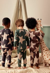 Kinder tragen Schlafanzüge mit großen beige Sternmustern auf dunkelgrauen, grünen und braunen Stoffen. Neben ihnen steht ein weiches Spielzeuggiraffe.