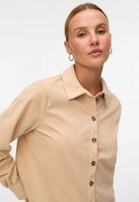 Camicia in corduroy a coste beige con colletto, chiusura a bottoni sul davanti e maniche lunghe. Presenta grandi bottoni marroni e una finitura testurizzata.