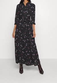 Zwart langemouwige jurk met bloemenpatronen in roze en paars, ceintuur in de taille en een zwierige rok. Gemaakt van een lichte, zachte stof.