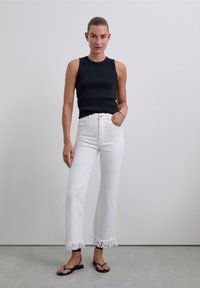 Kvinna står mot en enfärgad vägg och bär en ärmlös svart topp, vit högmidjade jeans med fransar, svarta sandaler och ett silverarmband.