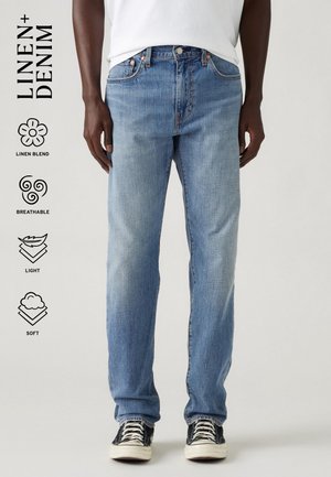 Personne portant un jean en mélange de lin bleu clair et des baskets basses noires avec lacets et semelles blanches sur un fond uni.