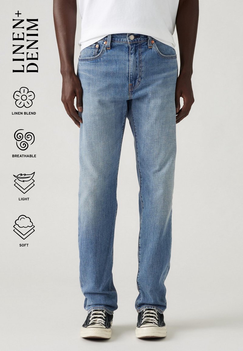 Personne portant un jean en mélange de lin bleu clair et des baskets basses noires avec lacets et semelles blanches sur un fond uni.