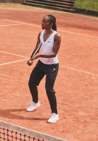 Sportists baltā bez piedurknēm tērpā un melnās biksēs, tur tennisu raketi uz māla laukuma. Baltas kurpes ar rozā akcentiem, sapītām matiem.