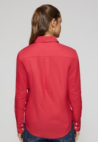 Camicia a maniche lunghe di un brillante rosso, realizzata in tessuto liscio. Presenta un colletto a punta, polsini con bottoni e un orlo arrotondato con una spallina posteriore.