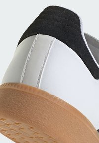 Białe trampki z czarnym zamszowym piętem, teksturowaną gumową podeszwą i kontrastującym przeszyciem. Posiadają górną część z gładkiej skóry i zaokrąglony kształt.