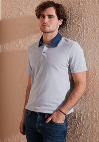 Světle šedé polo tričko s krátkým rukávem a džínovým límcem. Obsahuje tří-buttonový límec a jemně strukturovanou látku. Nosi se s modrými džíny.