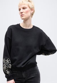 Άτομο με κοντά ξανθά μαλλιά που φοράει μαύρο φούτερ με λευκή κεντητή λουλουδένια διακόσμηση στα μανίκια και σκούρα τζιν, κοιτάζοντας προς τα πλάγια.