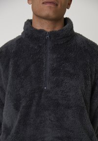Homme portant un pull en polaire à col montant gris foncé avec une demi-fermeture éclair, visible de la mâchoire jusqu'au milieu du torse, sur un fond uni.