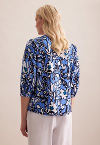 Blaue Bluse mit floralem Muster, puffärmel und abgerundetem Saum. Sie weist eine Mischung aus Marineblau, Hellblau und Weiß auf und ist aus leichtem Stoff gefertigt.