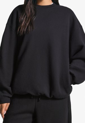 Frau trägt übergroßen schwarzen Sweatshirt und passende schwarze Hose, die Hände zum Teil in den Taschen, vor weißem Hintergrund.