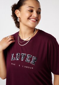 Bordeaux t-shirt met het woord "LOVER" in zwarte pailletten, met een ontspannen pasvorm, korte mouwen en een ronde halslijn. Inclusief zilveren gelaagde kettingen.