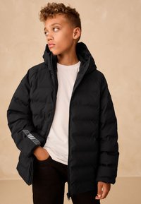 Schwarze Steppjacke mit Kapuze, mit gesteppter Struktur und einem grauen Logo am linken Ärmel. Getragen über einem weißen Hemd, kombiniert mit dunklen Hosen.