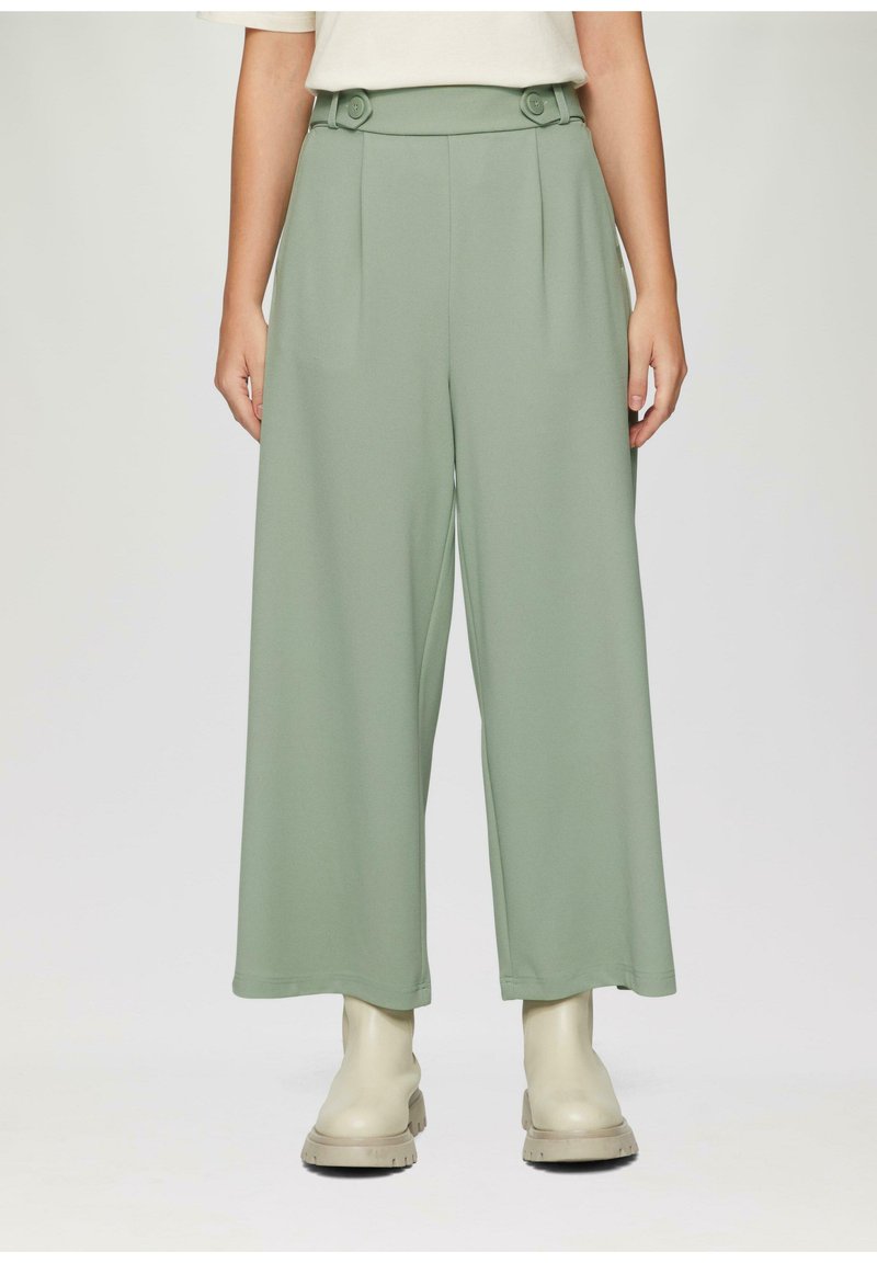Brede broek van zacht, lichtgroen stof. Kenmerkt zich door een hoge taille met dubbele knopen en voorplooien. Gepaard met beigerode enkellaarsjes.