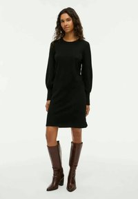 Frau mit lockigem Haar, die ein schwarzes, langärmeliges, knie-langes Kleid und braune, knielange, hochhackige Stiefel trägt, steht vor einem schlichten Hintergrund.