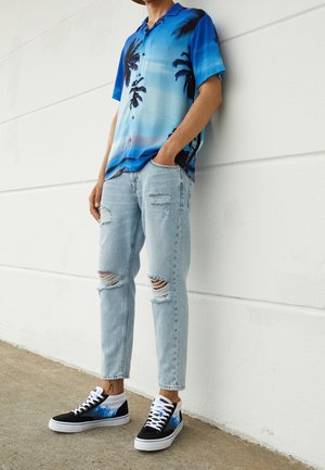 Osoba ubrana w niebieską tropikalną koszulę z krótkim rękawem, podarte jasnoniebieskie dżinsy i czarno-niebieskie wysokie trampki, stojąca przy białej ścianie.
