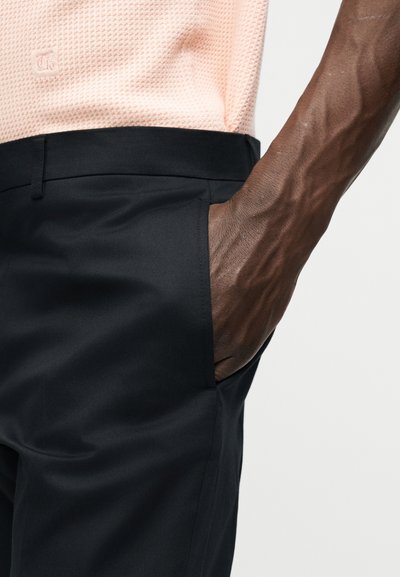 Σκούρου χρώματος δέρμα με εμφανείς φλέβες που αναπαύεται στην τσέπη μαύρου ραμμένου παντελονιού, φορώντας μια υφή ροζ ανοιχτό τοπ.