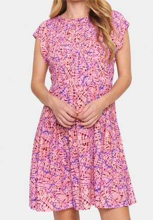 Nainen, joka käyttää lyhythihamaista, polvipituista vaaleanpunaista mekkoa, jossa on abstrakteja violetteja ja tummanpunaisia kuvioita, kädet ristissä edessä, pitkä vaalea tukka näkyvissä.