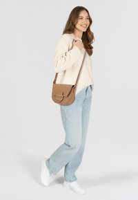 Braune Leder-Umhängetasche mit flacher Form, ausgestattet mit einem rechteckigen Deckel und einem runden Schnallenakzent. Kombiniert mit hellen Jeans und einem cremfarbenen Pullover.
