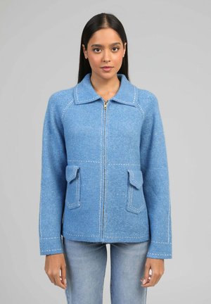 Jeune femme debout, visage tourné vers l'avant, portant une veste zippée bleu clair avec poches à rabat et un jean délavé, sur un fond gris uni.