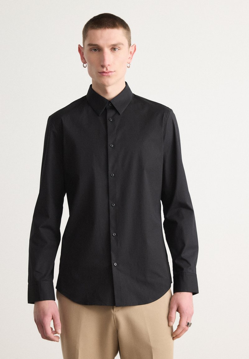 Camicia nera con bottoni, colletto, maniche lunghe e design aderente. Realizzata in tessuto liscio con un logo discreto sul petto.