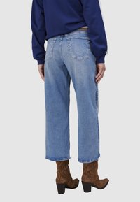 Jean bleu clair, taille haute, avec une coupe relax et une longueur raccourcie. Présente des poches arrière et un ourlet effiloché. Porté avec des bottes en suédine marron.