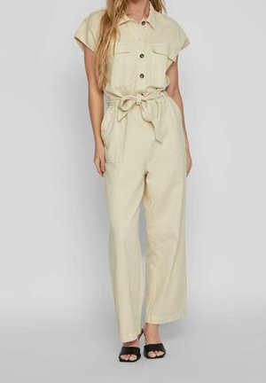 Kvinde iført beige, ærmeløs jumpsuit med knapper og bindebånd i taljen samt sorte åbne sandalhæle, stående foran en ensfarvet grå baggrund.