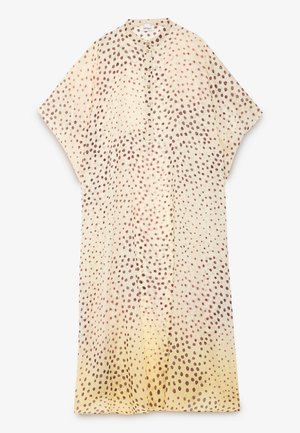 Kvinde iført en lysebeige bluse med mørkebrune prikker og en lang brun halskæde, stående mod en ensfarvet baggrund.