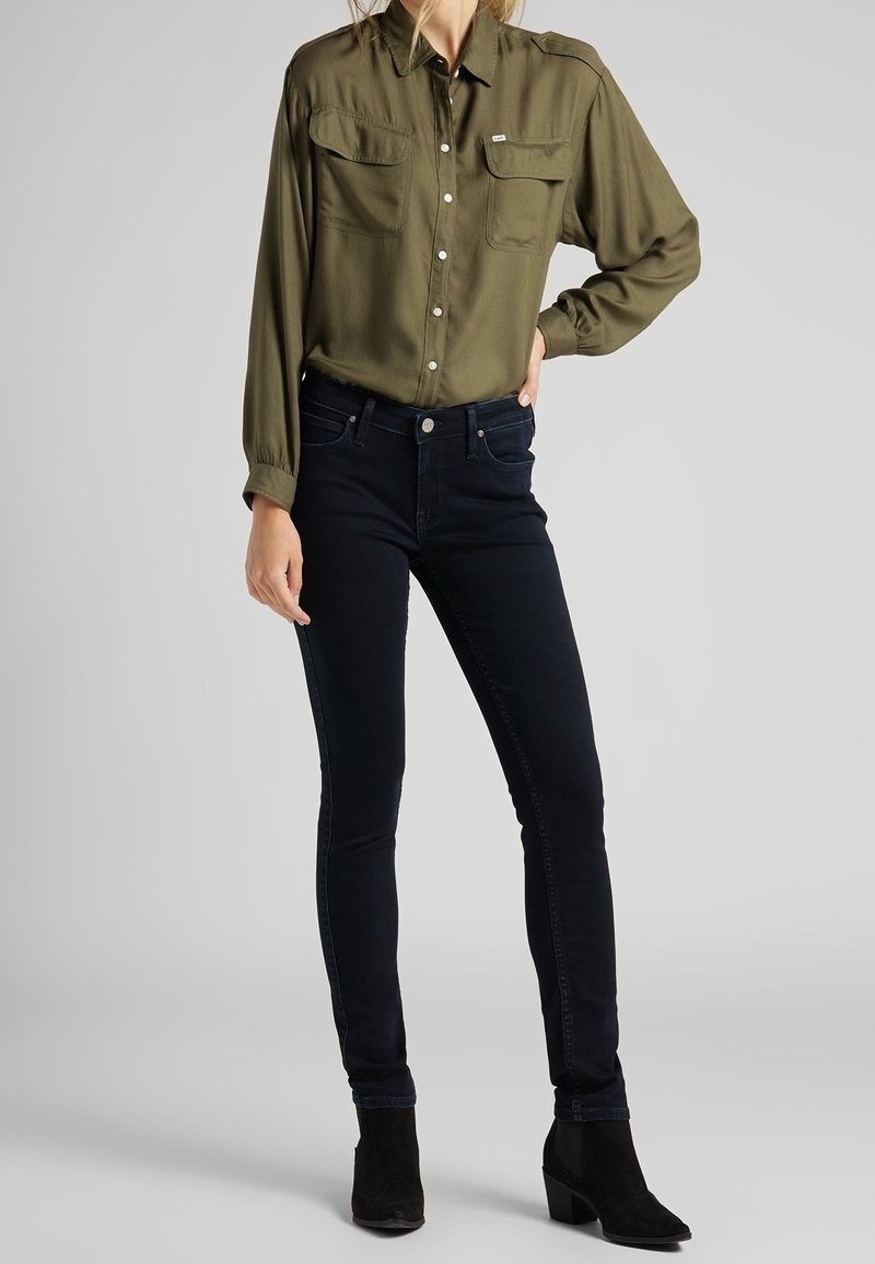 Camisa de manga larga en verde oliva con dos bolsillos en el pecho, combinada con unos jeans ajustados de color azul oscuro y botas negras hasta el tobillo. Tejido suave, corte estructurado.