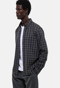 Chemise à carreaux noir et blanc avec un col boutonné, poche avant et manches longues, portée sur un t-shirt blanc et un pantalon gris.